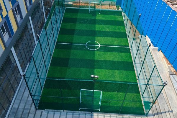 Aerial view of a small enclosed artificial turf futsal field with center circle, goals at both ends, and glass walls on the left and blue fencing on the right.