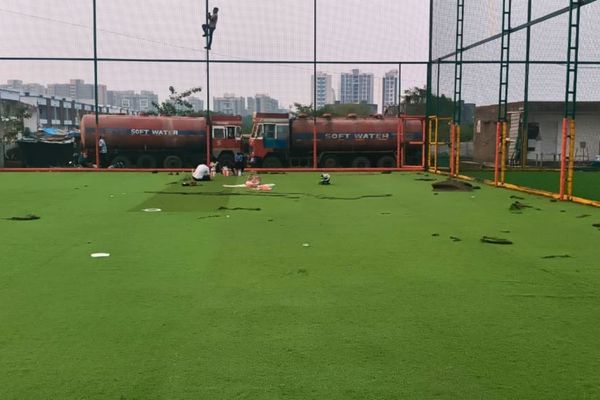 Rooftop soccer field with green turf, safety netting, and workers clearing debris; city skyline behind and a red water tanker labeled 'SOFT WATER' in the background.