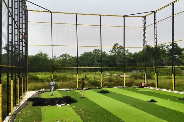 Outdoor cricket practice net enclosure with yellow-black framework and netting, players on green artificial turf.