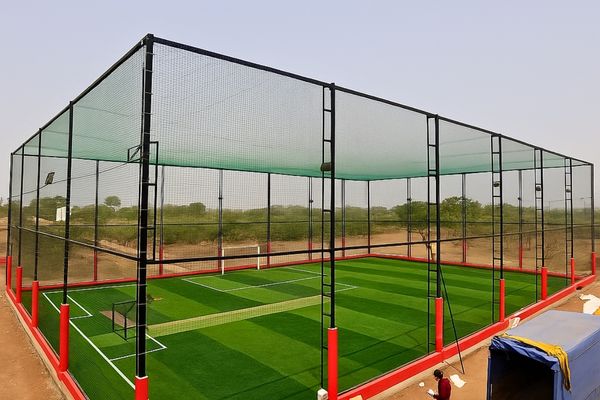 Cricket practice nets: a large fenced enclosure with net walls and an artificial turf pitch outdoors.